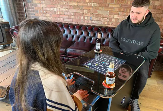 A man and a woman are seated at a glass-top cocktail table arcade machine playing Pacman in a brick-walled room. 